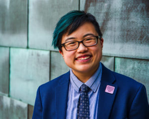 Lydia smiles and tilts their head slightly to the side, looking confidently at the camera. They are a young-ish East Asian person with a streak of teal in their short black hair, wearing glasses, a cobalt blue jacket and navy tie, with a blue copper wall behind them. Photo by Sarah Tundermann